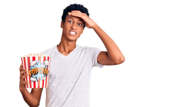 Young african american man holding popcorn stressed and frustrated with hand on head, surprised and angry face