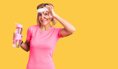 Young blonde woman wearing sportswear holding water bottle smiling happy doing ok sign with hand on eye looking through fingers