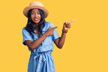 Young indian girl wearing summer hat pointing aside worried and nervous with both hands, concerned...