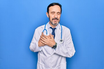 Middle age handsome doctor man wearing coat and stethoscope over isolated blue background smiling...