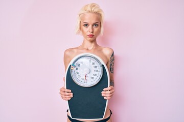 Young blonde woman with tattoo standing shirtless holding weighing machine relaxed with serious expression on face. simple and natural looking at the camera.