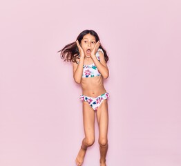 Adorable hispanic child girl on vacation wearing bikini scared with hands on face. Jumping over isolated pink background - obrazy, fototapety, plakaty