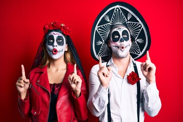 Couple wearing day of the dead costume over red amazed and surprised looking up and pointing with fingers and raised arms.