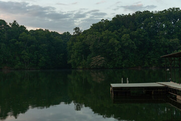 The swim platform on a dock at dawn with a ladder leading into the water surrounded by trees and their reflection in the water