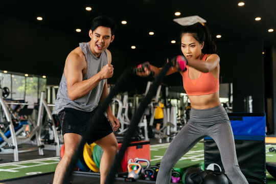 Asian Personal Trainer Coaching A Bodybuilding Woman To Perform Crossfit Battle Ropes Exercise In The Fitness Gym. Workout Training Coach Business Concept