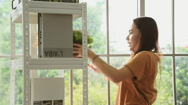 Young Asian woman in casual outfit doing chores at home using duster stick to clean book shelf