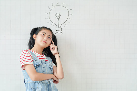 Pensive teenage Asian girl standing at white wall with light bulb drawing