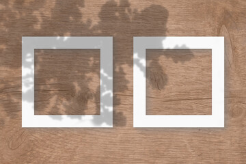 Two square sheet of white textured paper on the wooden table background. Mockup with an overlay of plant shadows. Natural light casts shadows from the oak leaves. Flat lay, top view