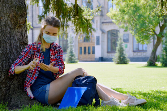 Young girl student in protective mask is sitting under tree near university and sanitize hands with antiseptic. Back to school after COVID-19 pandemic. New normal.