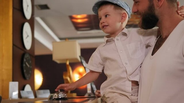 Close-up of a father and little son standing at the reception in the hotel lobby, a child playing with the hotel bell. Slow motion.