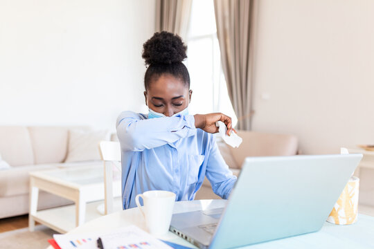 Young African businesswoman coughing into elbow in the home office. Businesswoman working in her home office while suffering from allergies. Women are sneezing and are cold during COVID-19 pandemic.