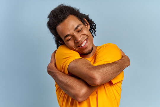 Handsome dark skinned young man in a yellow t-shirt hugs himself on a blue background. Guy with his eyes closed feels happy and positive, smiling confidently. Concept of selfishness and self-care.