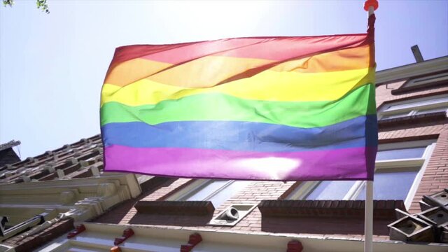 Slow motion: a rainbow flag waving with the facades of Amsterdam houses behind it on a sunny day.