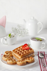 Breakfast. Fresh waffles on a white plate with strawberries and mint leaves on a white table. Fresh cottage cheese in a tea set.