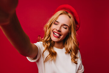 Wonderful white girl in beret making selfie with eyes closed and smiling. Portrait of adorable blonde female model with red lips.