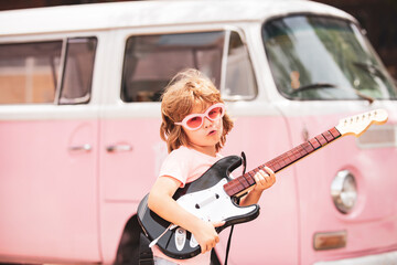 Kid musician playing the guitar like a rockstar on pink background in neon light. Cute pupil having...