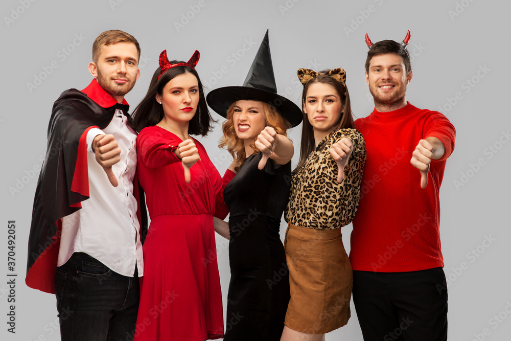 Wall mural friendship, holiday and people concept - group of friends in halloween costumes of vampire, witch, devil and cheetah showing thumbs down over grey background