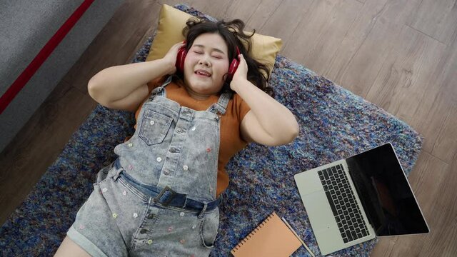 Asian plus size woman in headset lying on floor carpet listening and sing music, Working from home concept
