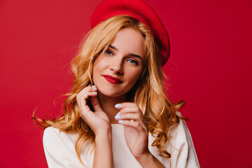 Emotional well-dressed woman with blonde hair looking to camera. Enchanting french girl standing on red background.