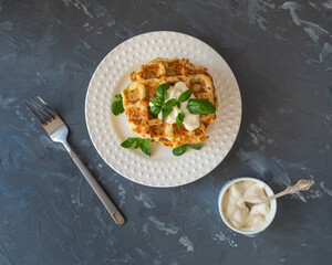 Vegetable waffles from young zucchini with cheese and basil on a white plateA pile of squash waffles, a white sauce bowl with creamy mushroom sauce on a gray background