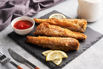 Fried fish hake Pollock with tomato sauce and slices of fresh lemon. Concept eating fatty fast food. Grey concrete background copy space.