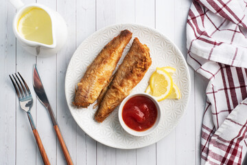Fried fish hake Pollock with tomato sauce and slices of fresh lemon. Concept eating fatty fast food. White wooden table with Copy space.