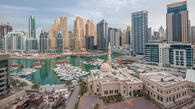 Yachts in Dubai Marina flanked by the Al Rahim Mosque and residential towers and skyscrapers aerial night to day transition timelapse. Modern high rise skyline with boats and wooden dhows