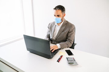 diagonal view of a white office table with a business worker working on a laptop with a face mask on with a calculator, cell phone, post-it, pens