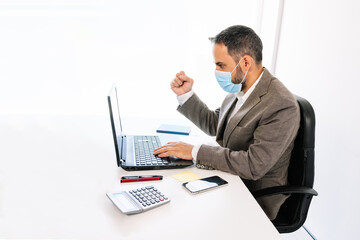 white office table with a business worker working and positively celebrating a new economic achievement with a face mask worn with a laptop, calculator, cell phone, post-it, pens, notebook