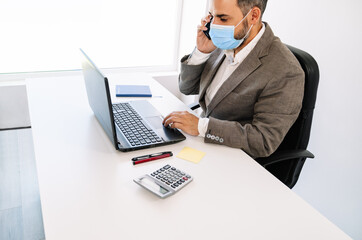 side view of a white office table with a business worker talking on a mobile phone with a face mask on with a laptop, calculator, mobile phone, post it, pens, notebook