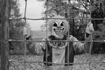  Halloween concept. Creepy clown costume.Evil clown on the playground in the autumn park. Black and white photo.Autumn holidays.Horror and fear.Carnival holidays in October
