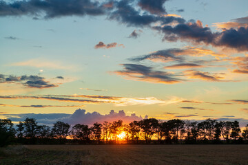 Fototapeta premium Magic sunset with sun low over the trees and dramatic clouds illuminated by last rays of sun.