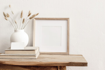 Square wooden frame mockup on vintage bench, table. Modern white ceramic vase with dry Lagurus ovatus grass, books and busines card. White wall background. Scandinavian interior.