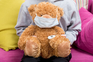Child holds fluffy toy bear with protective mask on his face and medical thermometer under his arm....