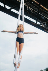 Child girl aerialist performs acrobatic tricks on aerial silk attached to the bridge support.