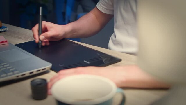 Close-up shot of young handsome man draws with a pen on a tablet at home at night at a table in the living room