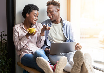 Joyful African American couple with laptop and coffee sitting on windowsill at cafe