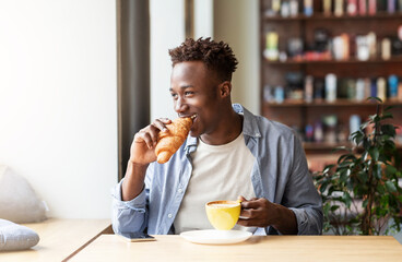 Funky African American guy having croissant with coffee for lunch at cafe