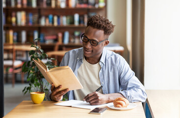 Cool black guy reading book and taking notes during his studies at cafe