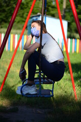 Young fresh cheerful hipster young woman sitting on a swing in the playground wearing stylish summer clothes. Warm evening tones. Mask. Coronavirus. Distance. Sunset.