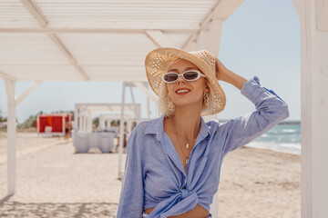 Outdoor summer fashion portrait of happy smiling woman wearing stylish straw hat, sunglasses, chain necklace, blue linen shirt posing on sea beach. Copy, empty space for text