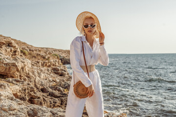 Outdoor summer fashion portrait of elegant woman wearing white linen suit, belt, straw hat, sunglasses, with round wicker bag, posing on rocks near sea. Copy, empty space for text - 370521008