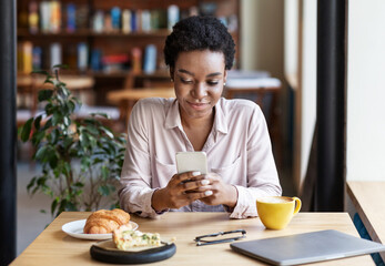 Young black woman using mobile phone for online communication at lunchtime in coffee shop
