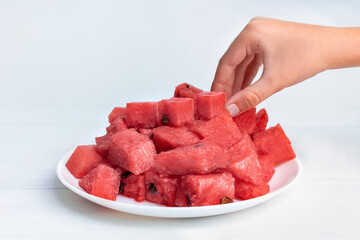 Child's hand takes juicy pieces of ripe red watermelon from a white plate. Seasonal sweet tropical fruit with seeds on the kitchen table, vitamins, summer time, portions of healthy dessert.