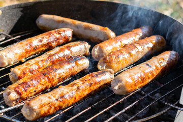 BBQ sausages are cooked on the grill in the meadow in the park on a sunny autumn day, closeup. Delicious juicy hot meat meal with smoky, backyard weekend barbeque picnic, family holiday meal.
