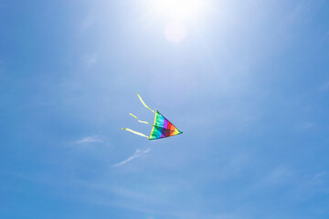 A kite in the shape of a delta and a rainbow color flies in the clear blue sky in the rays of the...