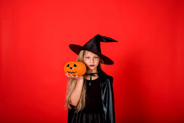 portrait of a serious girl in a witch costume in a hat with a pumpkin on a red background with a copy of space