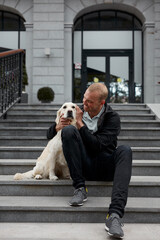 portrait of happy young caucasian guy sitting on stairs with golden retriever, hugs him, have fun...
