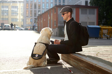 side view on handsome guy sitting outdoors, playing with dog, blind man have rest in city with...
