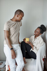 good-looking afro couple have rest at home, woman sits with yellow mug in hands, man talk with her. isolated in light room